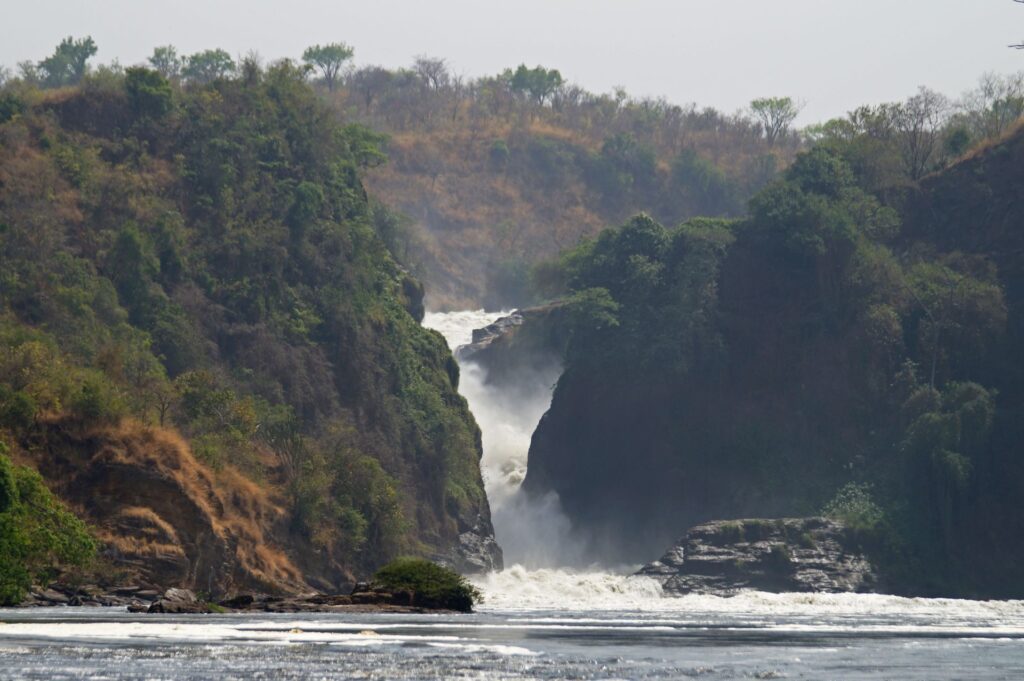 A distant view of a powerful waterfall between lush, forested hills with a mist rising from where the water crashes down.